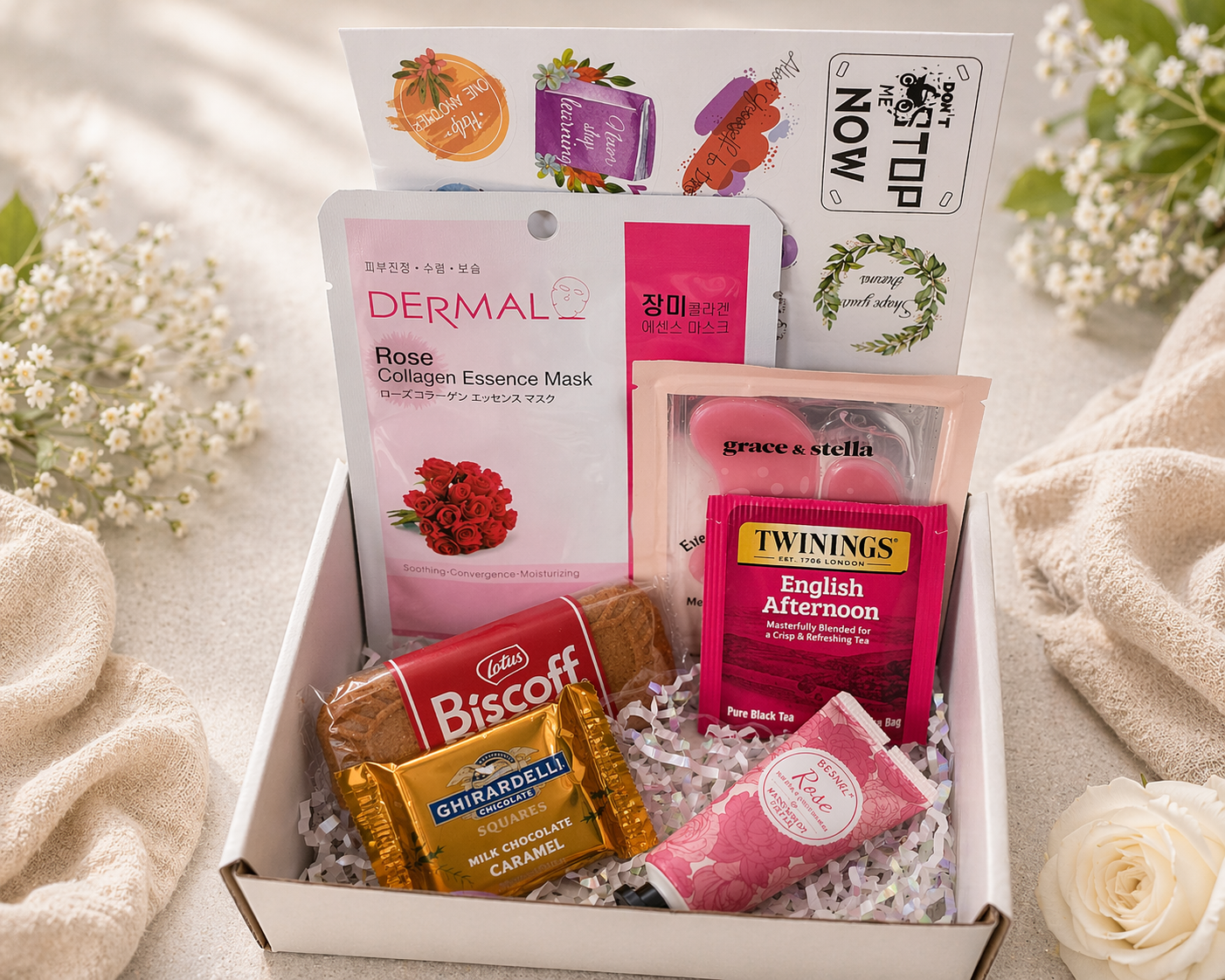 Assorted products including a Dermal rose collagen essence mask, Twining's English Afternoon tea, and Biscoff cookies in a box with flowers around.
