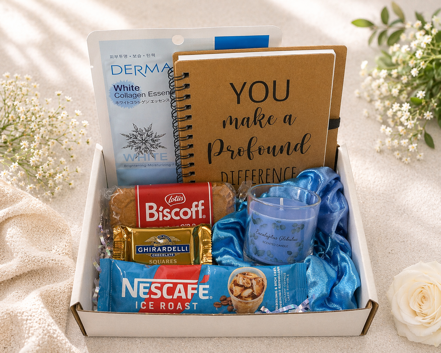 Gift box with snacks, a notebook, and a candle on a soft surface with flowers.