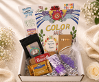 Gift box with 'Color Me' book, snacks, and decorative items on a soft background.