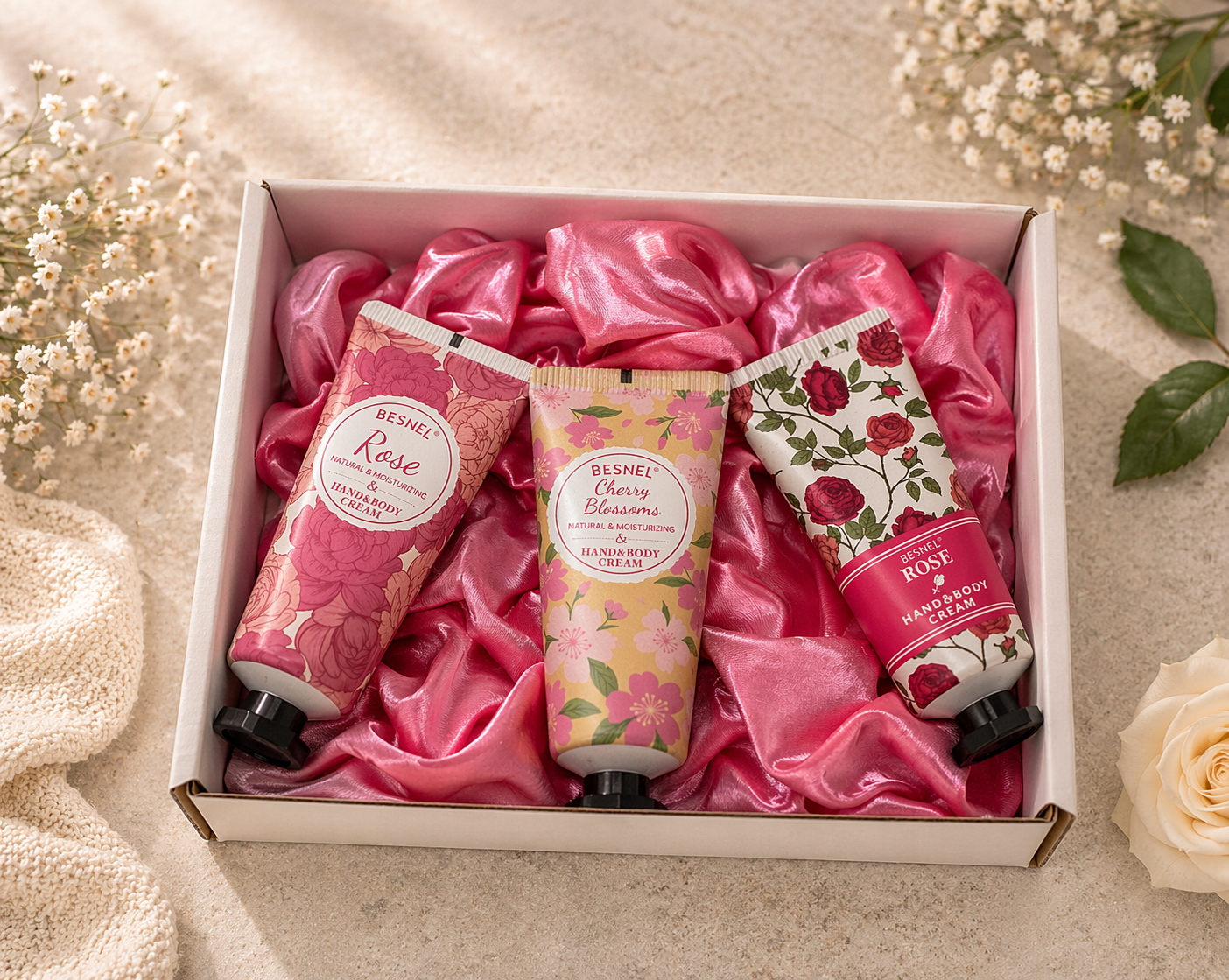 Three hand cream bottles in a box with pink satin lining, surrounded by flowers on a light background.