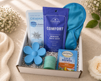 Gift box with bath products including a blue flower-shaped bar of soap, a packet of tea, and a bottle of bath soak on a beige background.