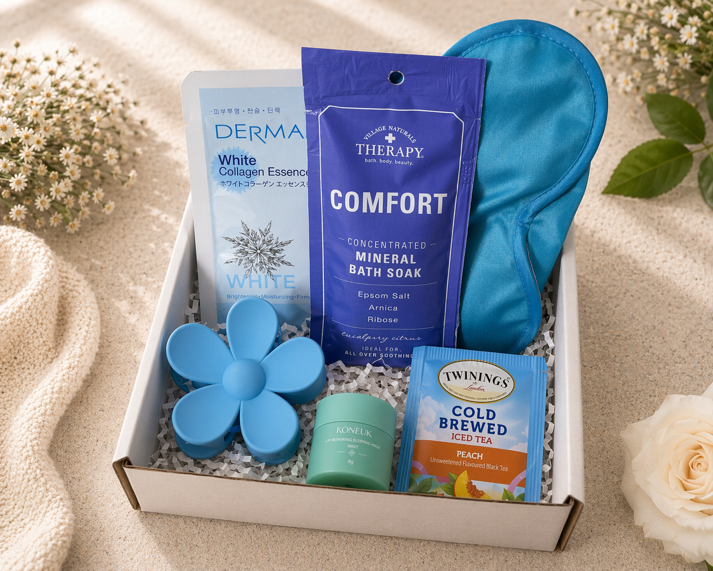 Gift box with bath products including a blue flower-shaped bar of soap, a packet of tea, and a bottle of bath soak on a beige background.