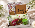 Gift box with a variety of items including a notebook, candles, and snacks on a light background.