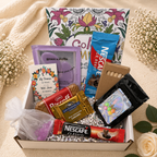 Gift box with snacks, Nescafe packets, and a card on a beige surface with flowers.