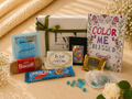 Gift box with snacks, a book titled 'Color Me Blessed', and decorative items on a light surface.